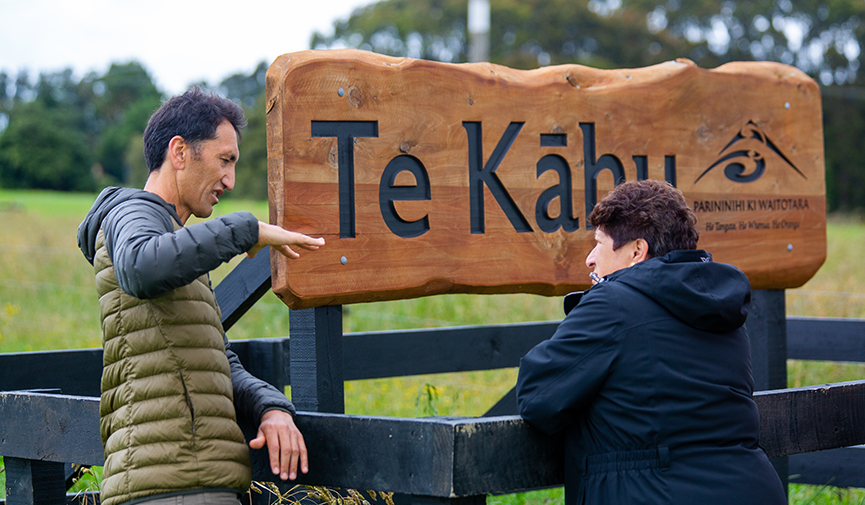 2 people standing talking in front of the Te Kāhu farm sign.