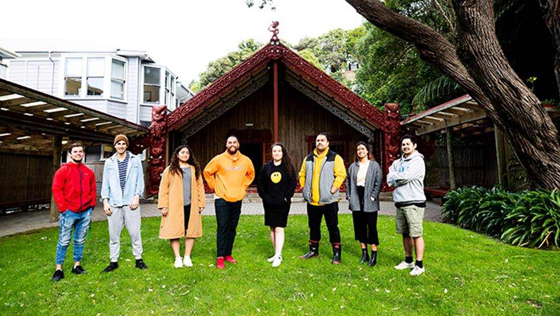 A group of people standing on the grass in front of the endtrance to a marae.