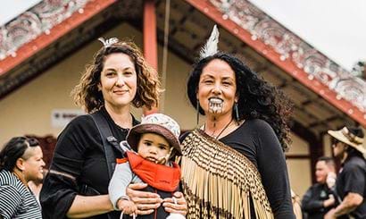 Two women and a baby in front of a marae.